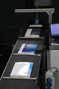 Boeken op een transportband in een industriële omgeving, tijdens een geautomatiseerd productieproces met een machine en beeldscherm op de achtergrond.